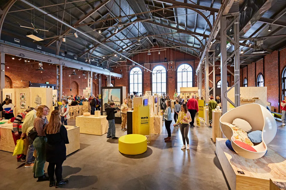 Eine Industriehalle mit Holzblöcken und Ausstellungsstücken, dazwischen Menschen und ein Schild mit der Aufschrift Werkschau Made in Sachsen